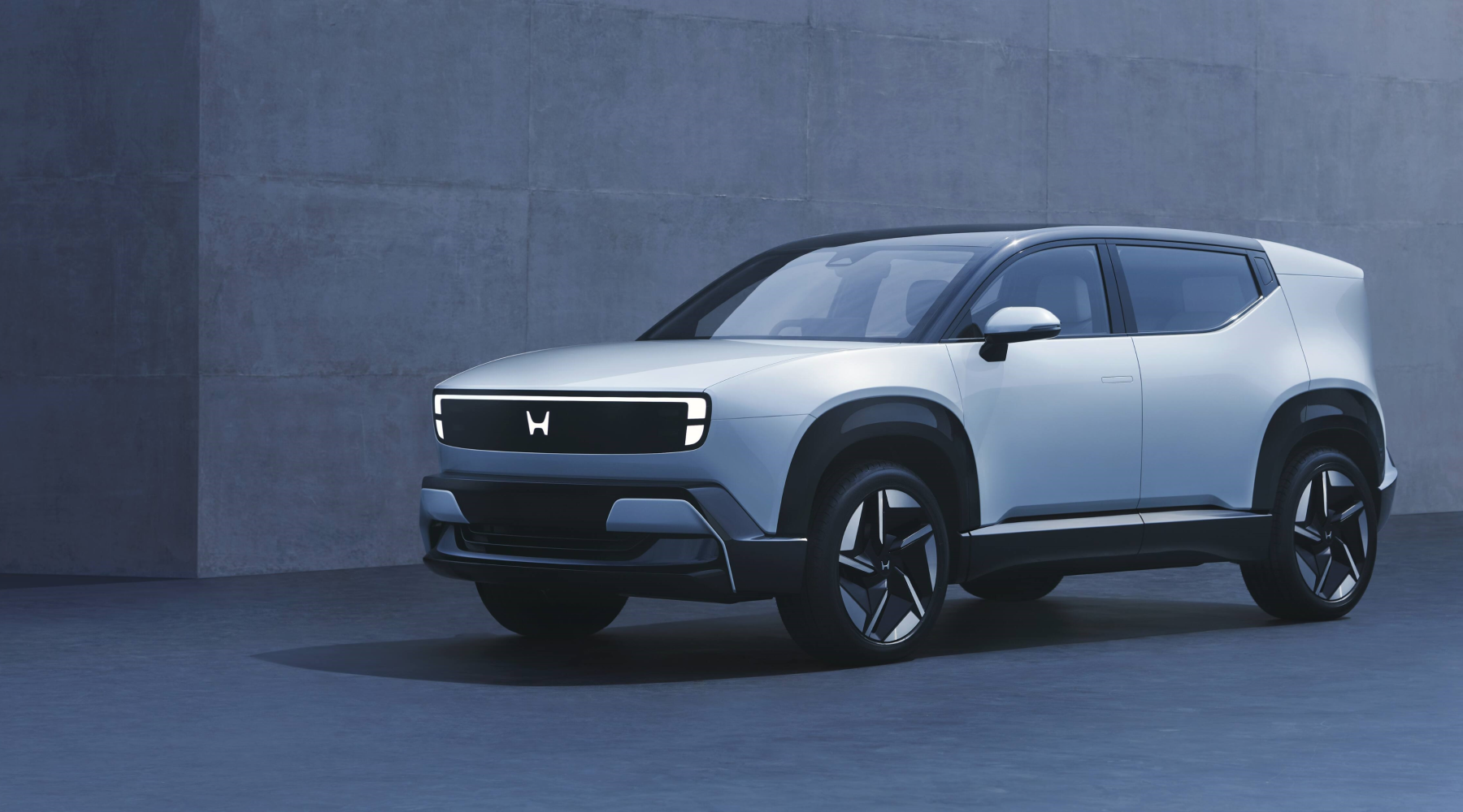 A sleek, modern Honda electric SUV concept in light blue, featuring a minimalist design with a distinctive front grille displaying the Honda logo. The vehicle is positioned against a gray concrete wall background.